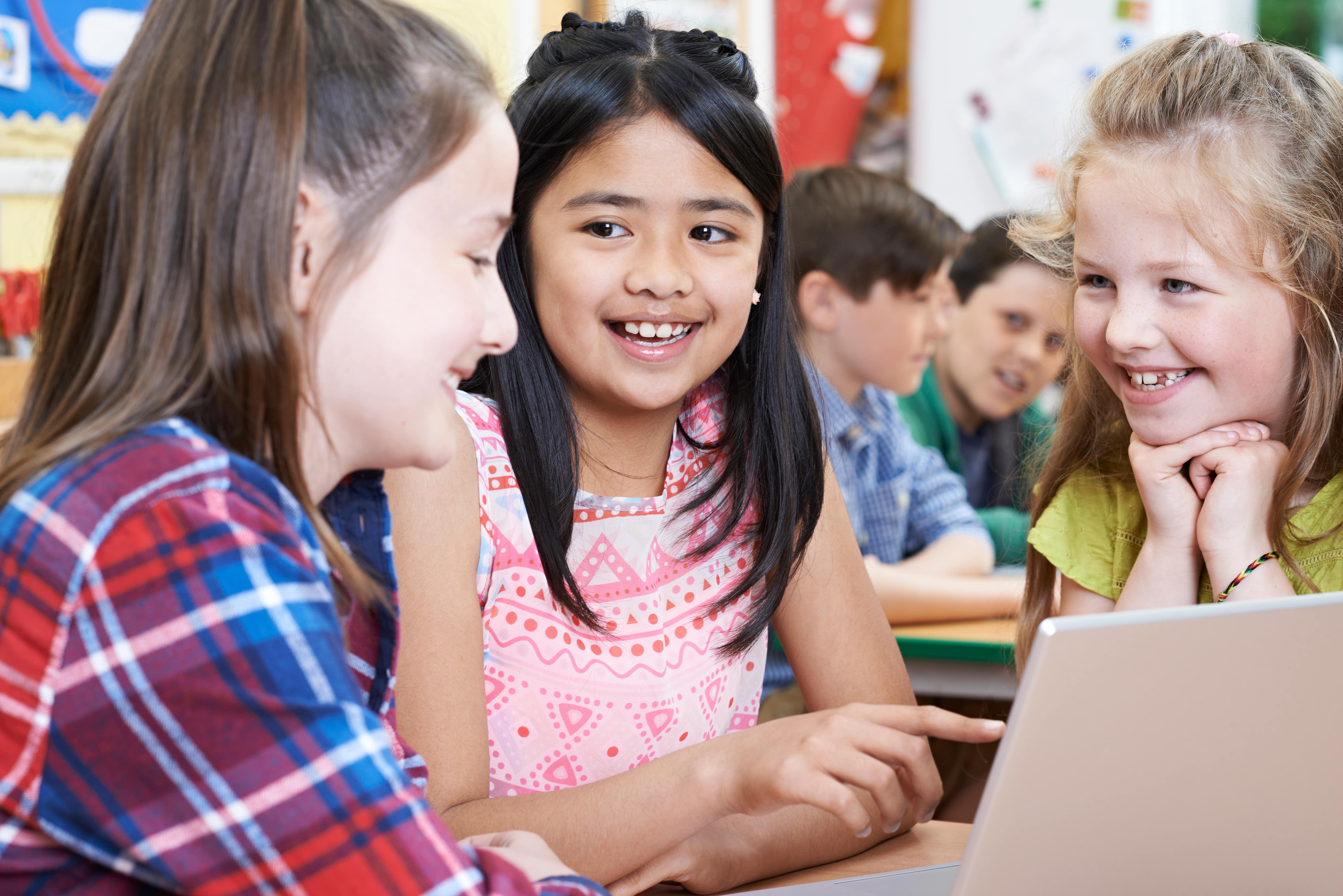 Three smiling elementary school aged girls sit together at a laptop