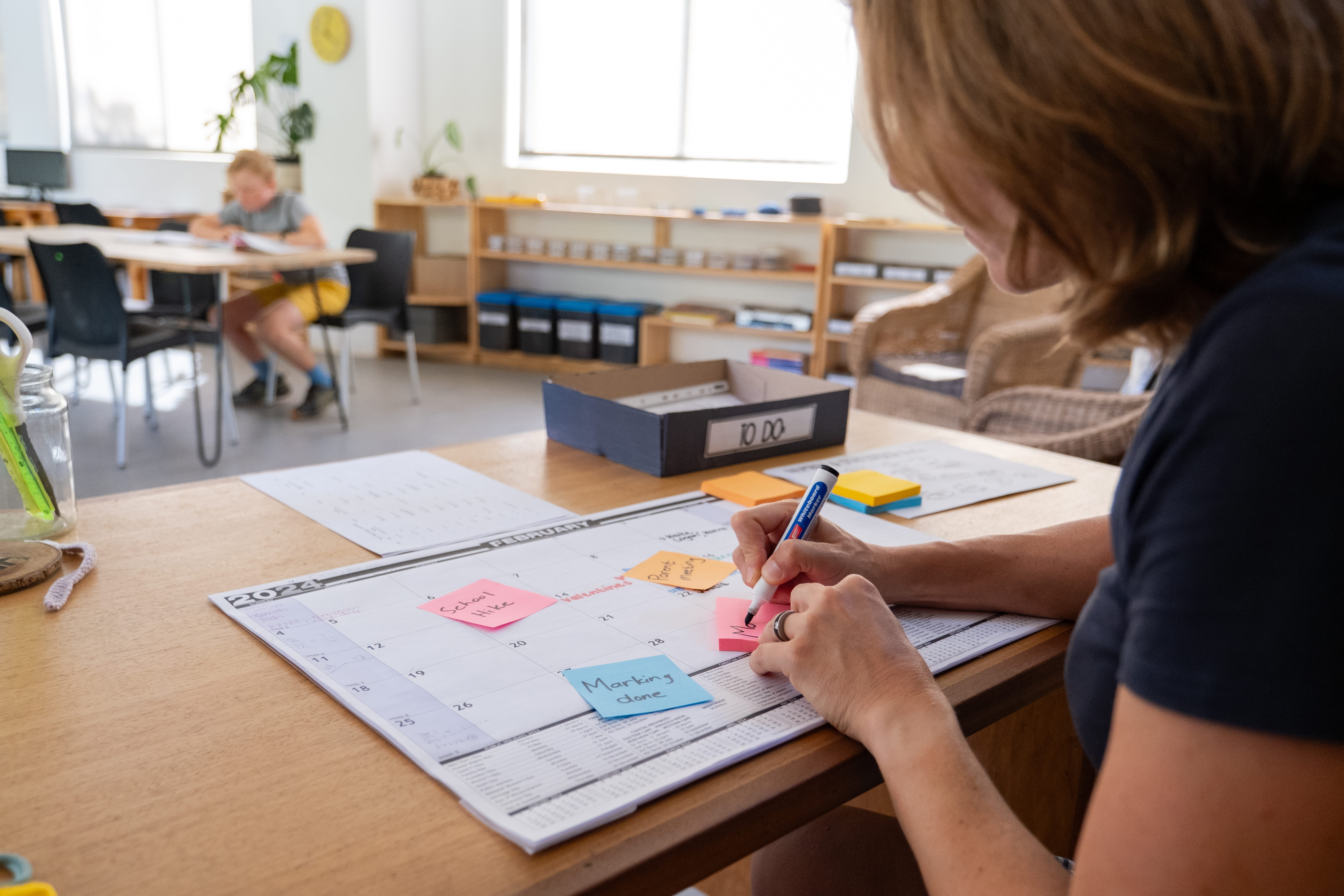 Female teacher sitting at desk in elementary school classroom using sticky notes for lesson planning.