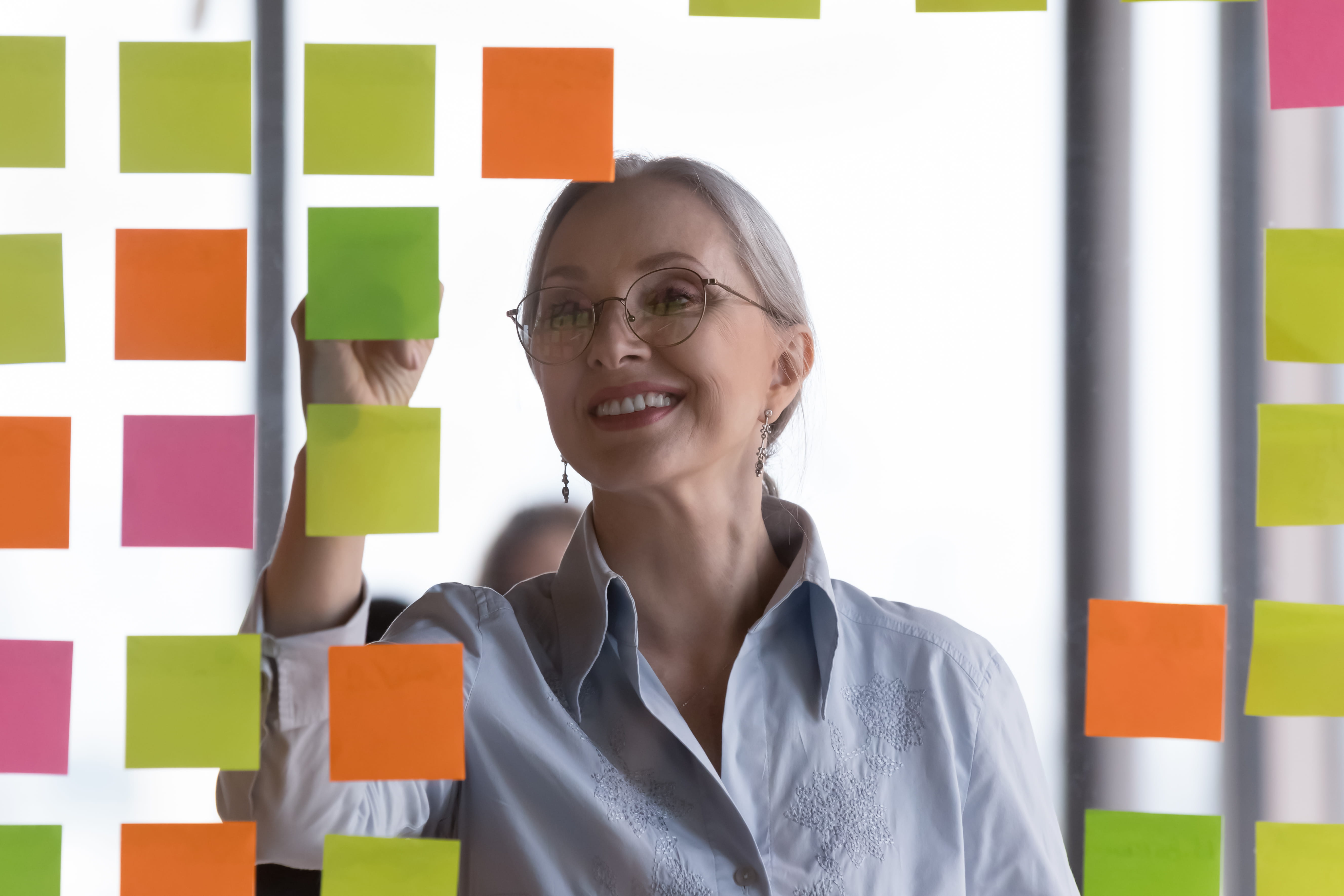 Older female teacher engaging in lesson design by writing on a grid of multicolored sticky notes.